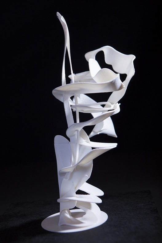 Es ist eine schlanke Skulptur zu sehen, die aus zwei ineinander geschlungenen Bändern besteht. Die Bänder repräsentieren in ihrer Form, Ausrichtung und Lage die Spur der Handbewegung beim Gestikulieren.